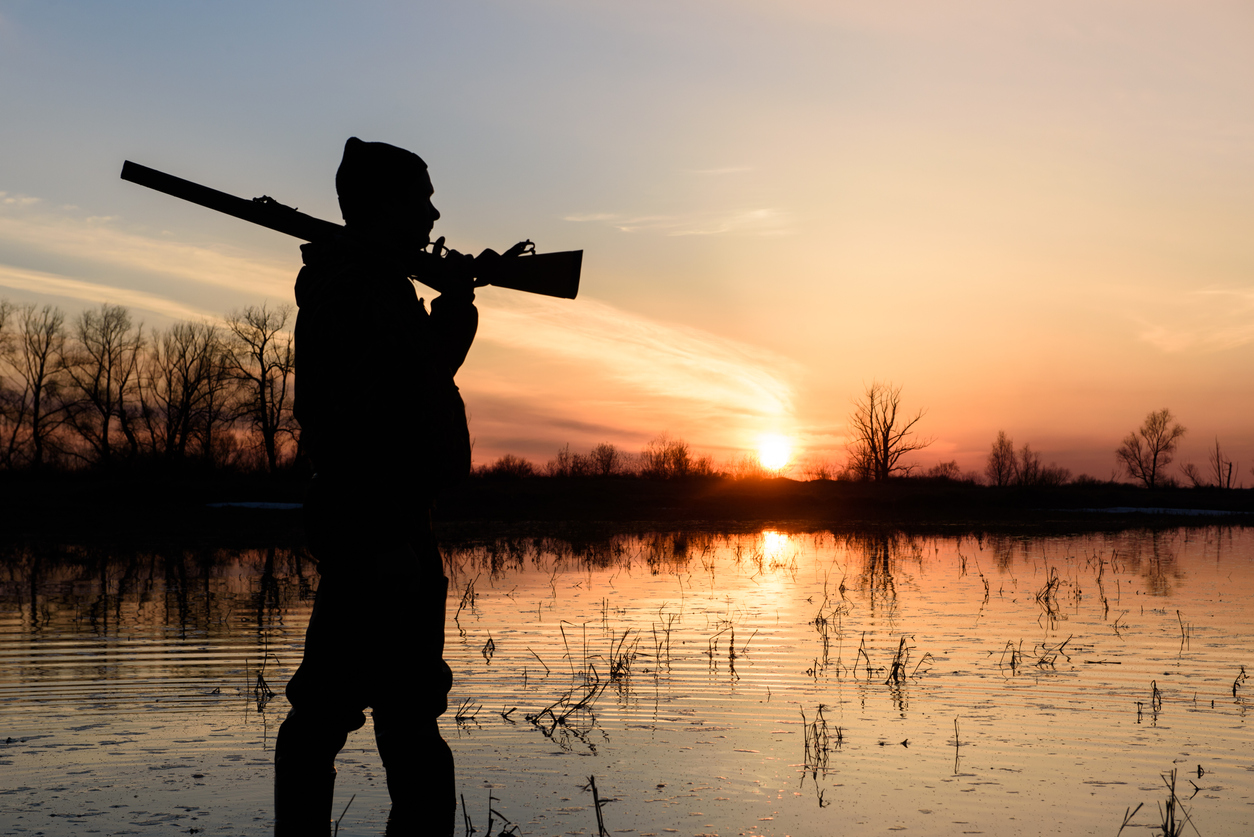 A hunter holding a gun while walking across the water silhouetted by the sunset