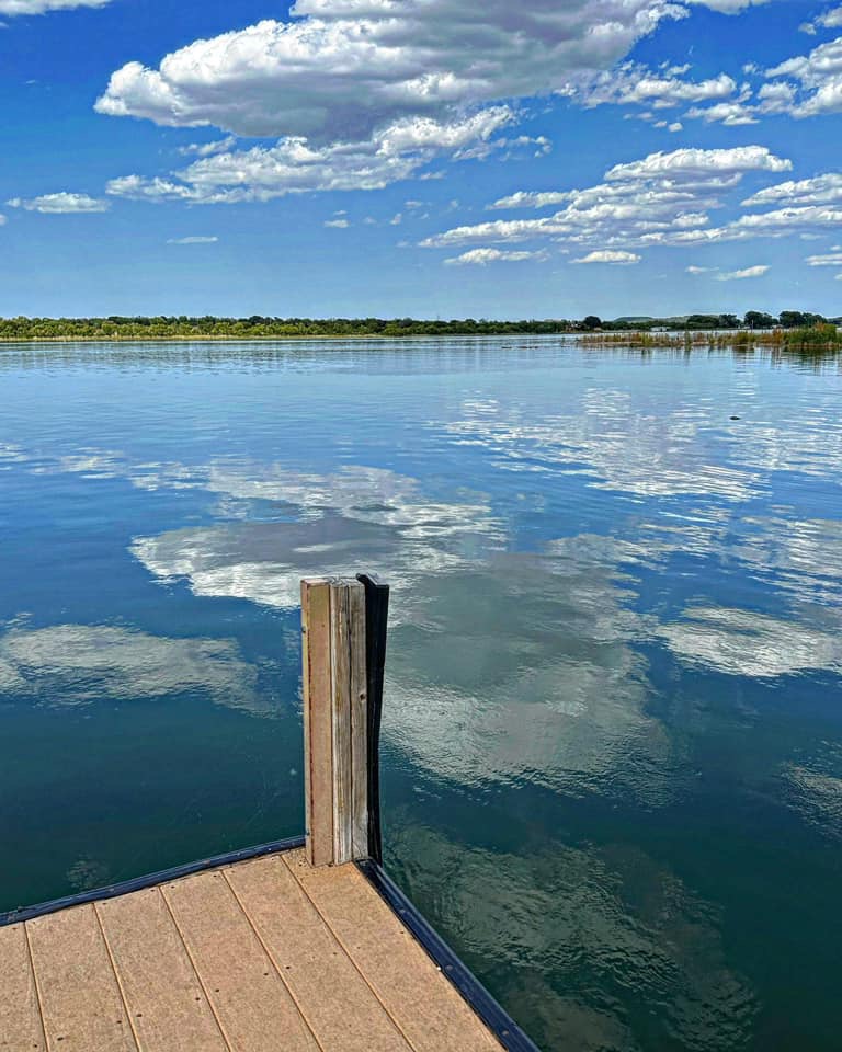View of a dock on the water which is reflecting the big white clouds